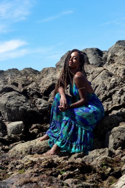 Mavi elbiseli, örgülü saçlı, kumsalın karanlık kayalarında oturmuş mavi gökyüzüne karşı güneşlenen güzel bir kadın..