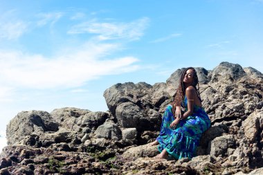 Mavi elbiseli, örgülü saçlı, kumsalın karanlık kayalarında oturmuş mavi gökyüzüne karşı güneşlenen güzel bir kadın..