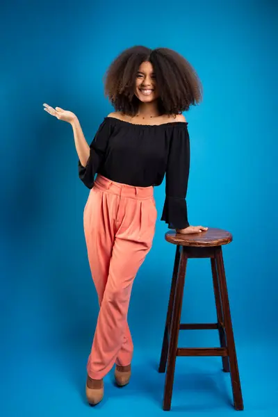 Young woman with black power hair, leaning on a wooden stool with her palm facing up. Isolated on blue background.