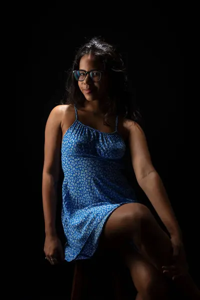 Young woman with straight hair wearing glasses and a blue dress posing for a photo. Isolated on black background.