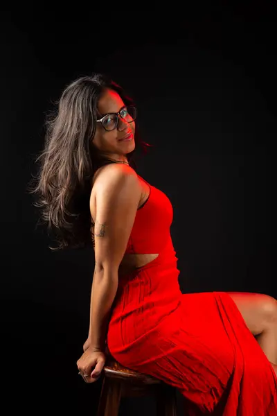 Beautiful young woman wearing glasses wearing red color outfit sitting posing for photo. Isolated on black background.