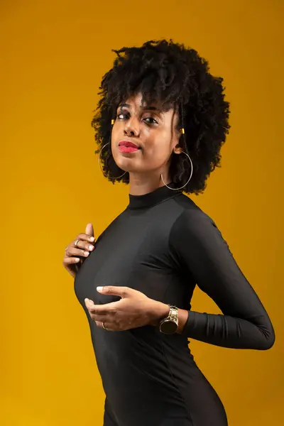 Young, sensual woman, with black power hair, standing, wearing black clothing and posing for a photo. Isolated on yellow background.