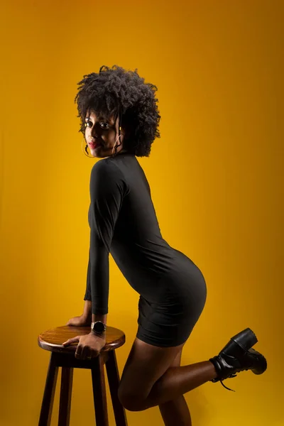 Sensual young woman with black power hair, standing, wearing black clothing, posing for a photo next to a wooden stool. Isolated on yellow background.