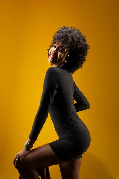 Sensual young woman with black power hair, standing, wearing black clothing, posing for a photo next to a wooden stool. Isolated on yellow background.