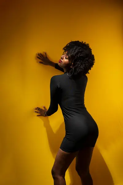 Young sensual woman with black power hair, wearing black clothes leaning against a yellow wall posing for a photo.