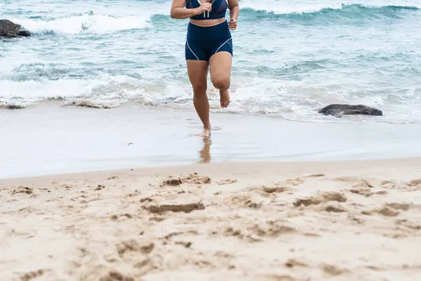Lower body of a young fitness woman running on the beach sand towards the camera. Healthy lifestyle.