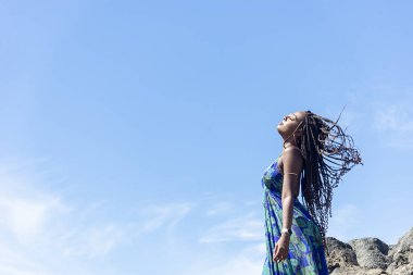 Mavi elbiseli güzel bir kadın sahildeki kayalıklarda dikilip mavi gökyüzüne dokunuyor..