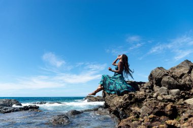 Mavi elbiseli ve örgülü saçlı güzel bir kadın kumsalın kenarında oturmuş mavi gökyüzüne karşı güneşleniyor..