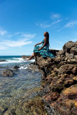 Mavi elbiseli ve örgülü saçlı güzel bir kadın kumsalın kenarında oturmuş mavi gökyüzüne karşı güneşleniyor..