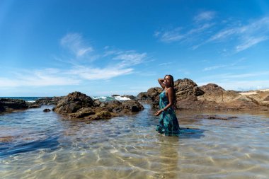 Mavi elbiseli ve örgülü saçlı güzel bir kadın sahilde suyun üzerinde duruyor mavi gökyüzüne karşı poz veriyor..