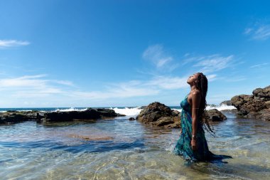 Mavi elbiseli, saçları örgülü, kumsalda duran, yürüyen ve mavi gökyüzüne bakan güzel bir kadın..