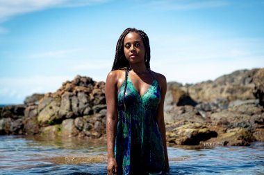 Sahil suyunda mavi elbiseli ve örgülü güzel bir kadın kameraya doğru yürüyor arkadaki kayalara ve mavi gökyüzüne doğru.