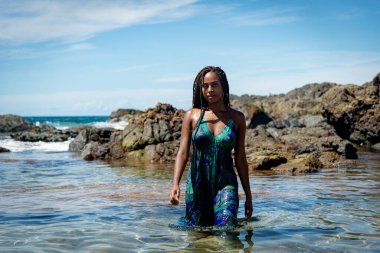 Sahil suyunda mavi elbiseli ve örgülü güzel bir kadın kameraya doğru yürüyor arkadaki kayalara ve mavi gökyüzüne doğru.