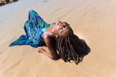 Mavi elbiseli, saçları örgülü, sırtı kapalı bir şekilde kumsalda oturan güzel bir kadın. Seyahat eden mutlu kişi.