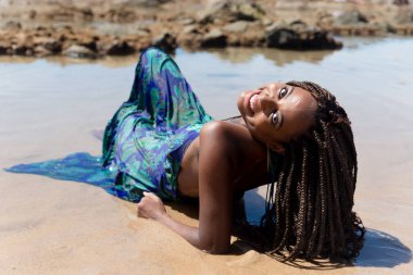 Mavi elbiseli ve örgülü saçlı güzel bir kadın kumsalda oturuyor ve sırtı kameraya bakıyor. Seyahat eden mutlu kişi.