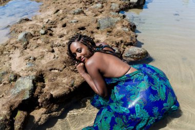 Saçları örgülü güzel bir kadın sahilde kayalara yaslanıyor ve fotoğraf için poz veriyor. Seyahat eden mutlu kişi.