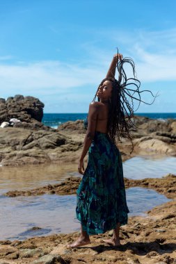 Güzel bir kadının portresi, kumsaldaki kayaların tepesinde mavi elbiseler giyip örgülü saçlarına dokunuyor. Seyahat eden mutlu kişi.