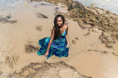 Saçları örgülü güzel bir kadın kumsalda uzun mavi bir elbiseyle güneşleniyor. Seyahat eden mutlu kişi.