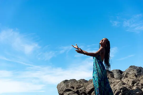 Beautiful woman, in blue clothes and braided hair, standing on the dark rocks of the beach with open arms against blue sky.