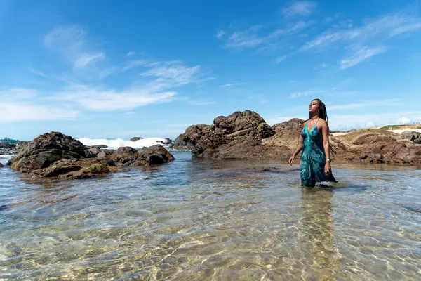 Mavi elbiseli, saçları örgülü, kumsalda duran, yürüyen ve mavi gökyüzüne bakan güzel bir kadın..