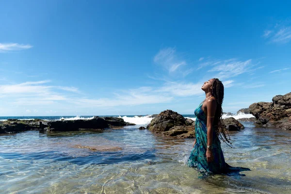 Mavi elbiseli, saçları örgülü, kumsalda duran, yürüyen ve mavi gökyüzüne bakan güzel bir kadın..