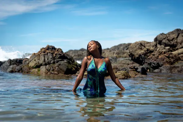 Mavi elbiseli güzel bir kadın ve kumsalda saçları örgülü bir kadın. Arka planda kaya ve mavi gökyüzüne karşı fotoğraf için poz veriyor..