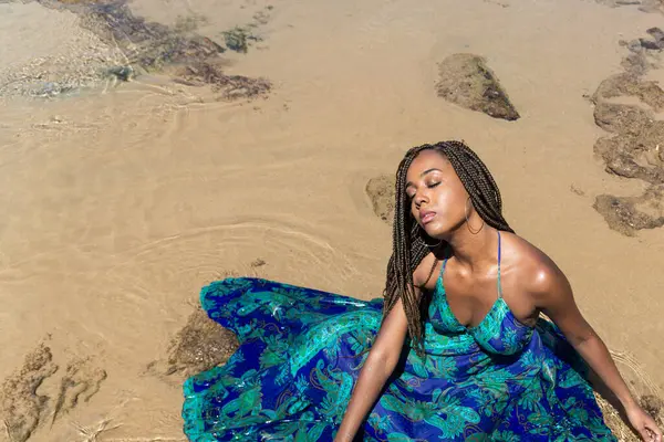 Saçları örgülü güzel bir kadın kumsalda uzun mavi bir elbiseyle güneşleniyor. Seyahat eden mutlu kişi.