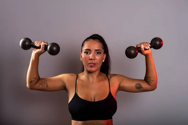 Beautiful brunette woman, in gym clothes, holding black dumbbells at head height. Healthy life. Against gray background.