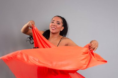 Siyah elbiseli esmer kadın gri arka planda turuncu bir bezle jimnastik yapıyor..