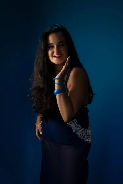 Portrait of beautiful woman with black hair, cheerful, confident, in a blue dress, looking at the camera with her hand on her chin. Isolated on dark blue background.