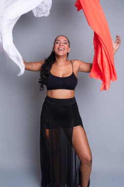 Siyah elbiseli esmer kadın gri arka planda beyaz ve turuncu elbiseli jimnastik hareketler yapıyor..