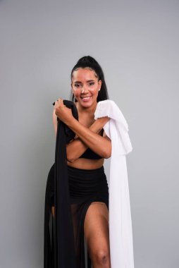 Siyah elbiseli esmer kadın gri arka planda beyaz ve siyah giysiler tutuyor..