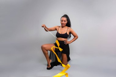 Jimnastikçi kadın, diz çöküp sarı kurdeleyle hareket ediyor. Gri arkaplanda izole.