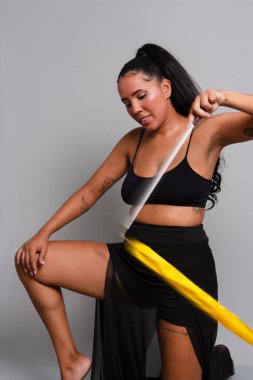 Jimnastikçi kadın, yerde diz çöküp sarı kurdeleyle dans ediyor. Gri arkaplanda izole.