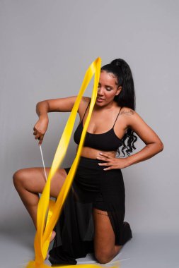Jimnastikçi kadın, yerde diz çöküp sarı kurdeleyle dans ediyor. Gri arkaplanda izole.