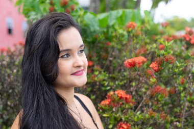 Güzel bir genç kadın portresi, uzun siyah saçlı, arka plandaki çiçeklere karşı sol tarafa bakıyor. Seyahat eden kişi.