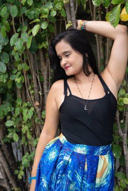 Güzel bir genç kadın, uzun siyah saçlı, elleri arkasında yeşil bir bitkiye yaslanmış. Seyahat eden kişi.