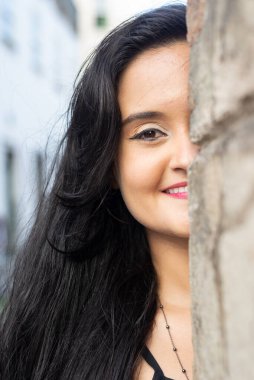 Güzel bir kadının yüzünün yarısının yakın plan portresi, siyah saçlı, dışarıda çimento duvara yaslanmış. Seyahat eden mutlu kişi.