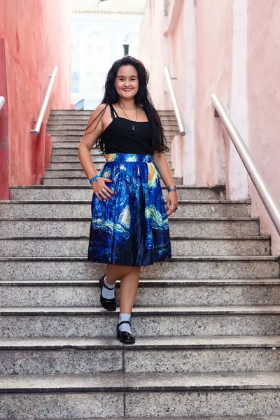Uzun siyah saçlı, siyah ve mavi elbiseli, merdivenlerden inip kameraya bakan güzel bir genç kadının portresi. Seyahat eden mutlu kişi.