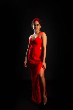 Salvador, Bahia, Brezilya - 09 Aralık 2023: güzel esmer kadın, reçeteli gözlük takıyor, kırmızı elbiseler giyiyor, ayakta. Siyah arkaplanda izole.