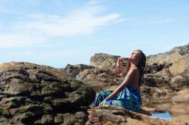 Salvador, Bahia, Brezilya - 10 Ocak 2024: Mavi elbiseli güzel bir kadın, elleri örgülü kayalıklarda oturuyor, kumsalda güneşleniyor. Mavi gökyüzüne karşı.