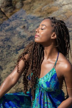 Salvador, Bahia, Brezilya - 10 Ocak 2024: Mavi örgülü ve mavi elbiseli güzel bir kadının portresi, kumsaldaki kayalarda mavi gökyüzüne karşı poz veriyor..