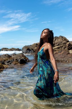 Salvador, Bahia, Brezilya - 10 Ocak 2024: Mavi elbiseli ve örgülü saçlı güzel bir kadın sahilde yürüyor ve mavi gökyüzüne karşı poz veriyor..