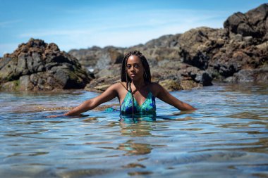 Salvador, Bahia, Brezilya - 10 Ocak 2024: Mavi elbiseli güzel bir kadın ve kumsal suyunda yarı gövdesi olan örgülü saçlar. Seyahat eden kişi.