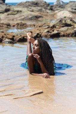 Salvador, Bahia, Brezilya - 10 Ocak 2024: Saçı örgülü ve mavi elbiseli güzel bir kadın, arka plandaki kayalara karşı poz veriyor..