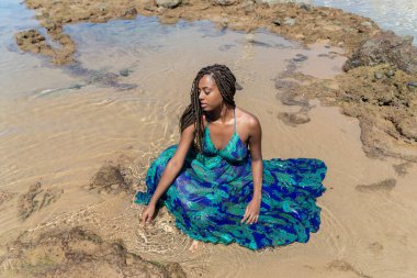 Salvador, Bahia, Brezilya - 10 Ocak 2024: örgülü saçlı güzel bir kadın kumsalda uzun mavi bir elbiseyle güneşleniyor. Seyahat eden mutlu kişi.