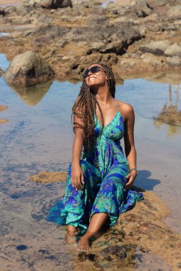 Güzel bir kadın, saç örgüleri ve mavi elbisesiyle kumsalda güneşlenen kayalarda oturuyor. Seyahat eden kişi.