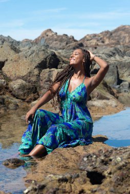 Güzel bir kadın, mavi elbiseli, elleri örgülü kayalıklarda oturuyor, kumsalda güneşleniyor. Seyahat eden kişi.