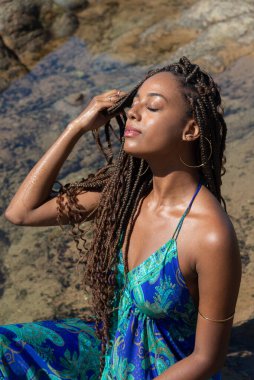 Mavi elbiseli güzel bir kadın kayalıklarda oturuyor, gözleri kapalı saçlarına dokunuyor, kumsalda güneşleniyor. Mavi gökyüzüne karşı.