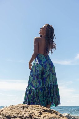Güzel bir kadın, saç örgüleri ve mavi elbiseleriyle sahildeki kayalıklarda dikilip mavi gökyüzüne karşı poz veriyor..